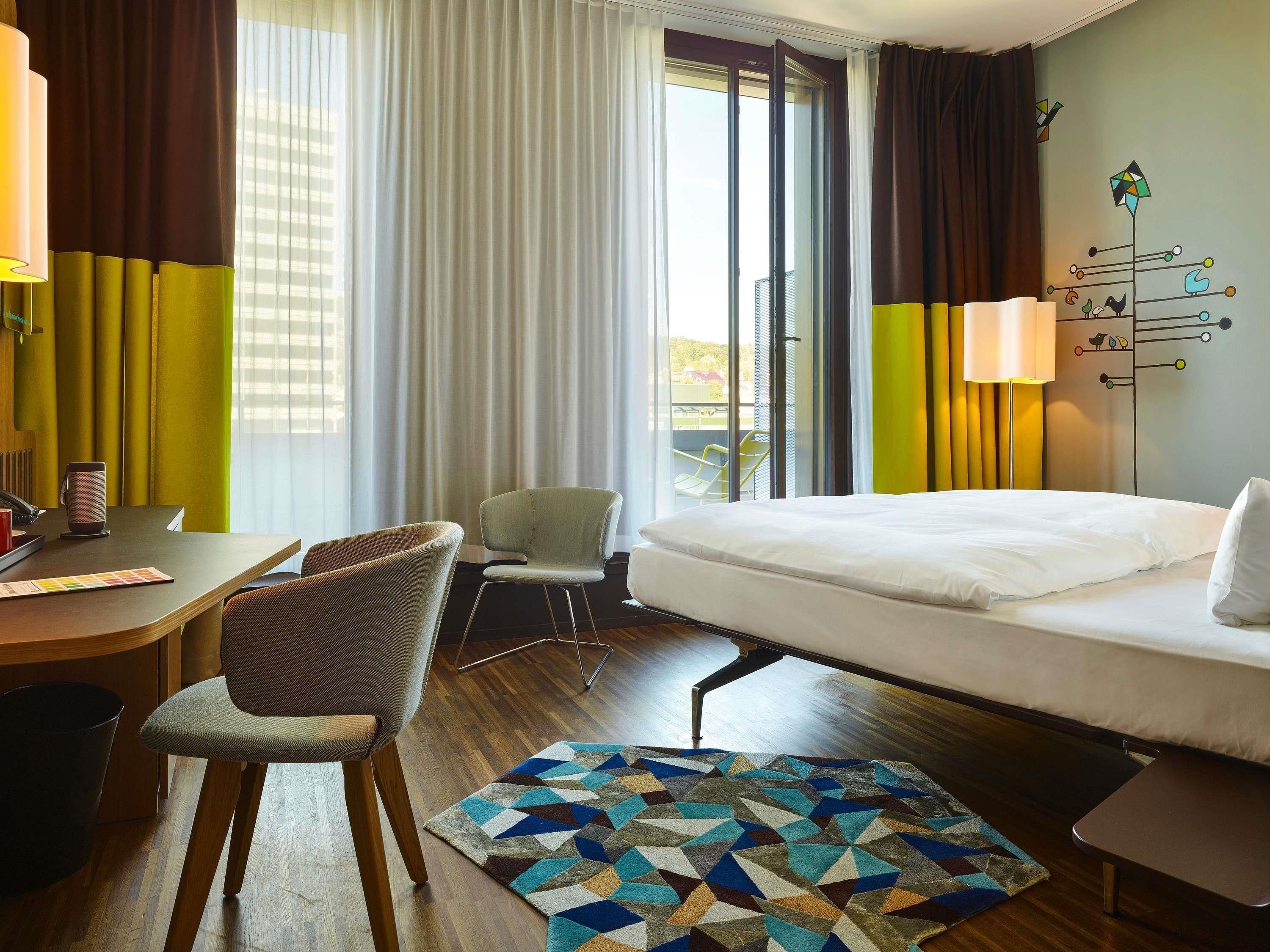 A hotel room with a bed, a desk with a chair, a coffee maker, and a window with curtains.