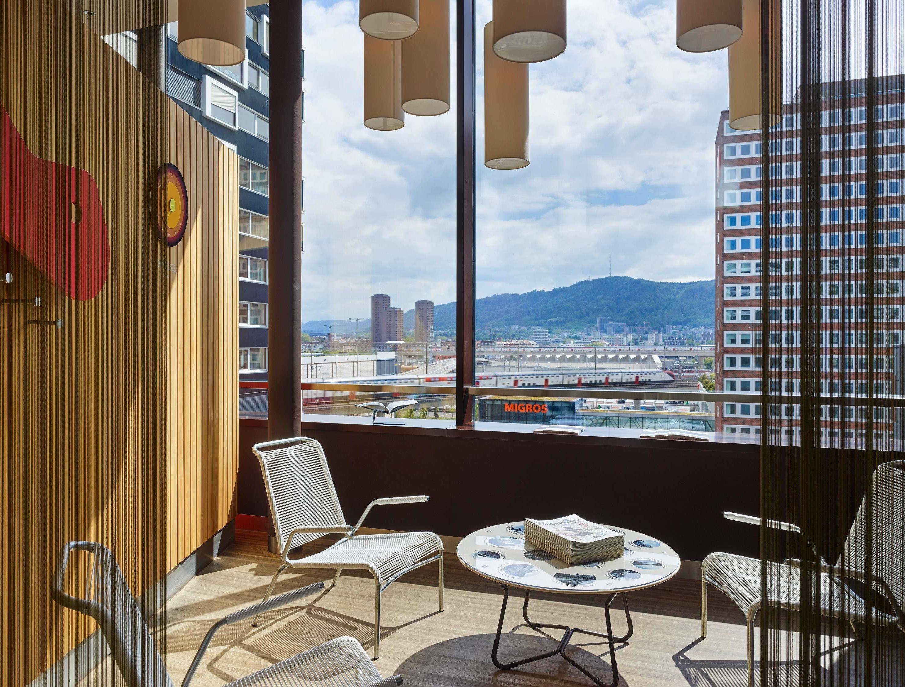 A modern hotel room with a large window offering a city view, a round table with chairs, and a magazine on the table.