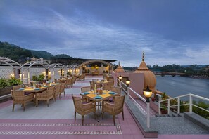 An outdoor dining area with tables and chairs, a view of a river, and a bridge in the background. - Ganga Lahari, Haridwar (Haridwar)