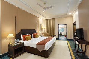 A hotel room with a large bed, a TV, a ceiling fan, and a view of the outdoors. - Ganga Lahari, Haridwar (Haridwar)