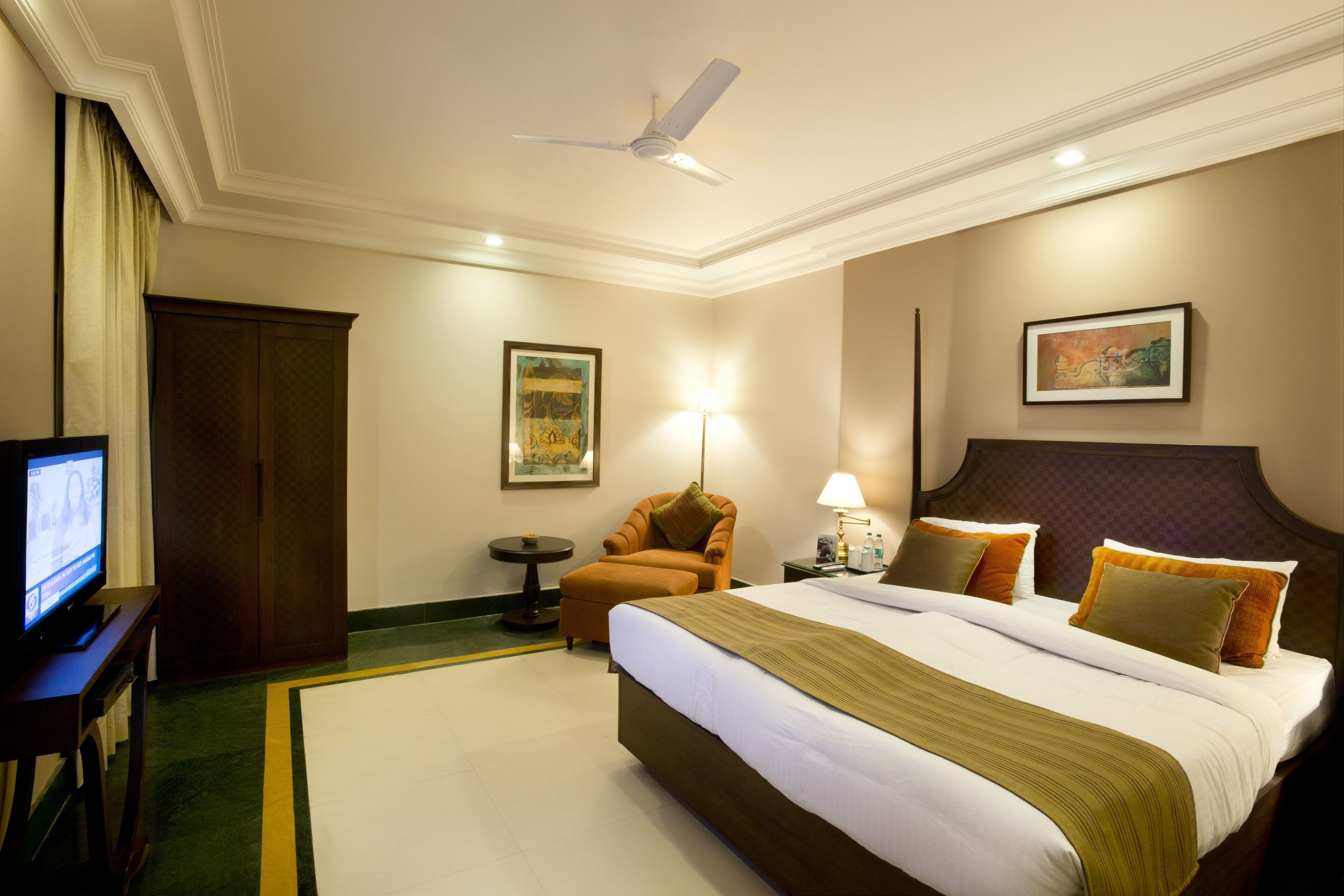 A hotel room with a bed, a television, a chair, a lamp, and artwork on the walls.