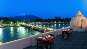 A terrace with a table set for dining, overlooking a river and a bridge. - Ganga Lahari, Haridwar (Haridwar)