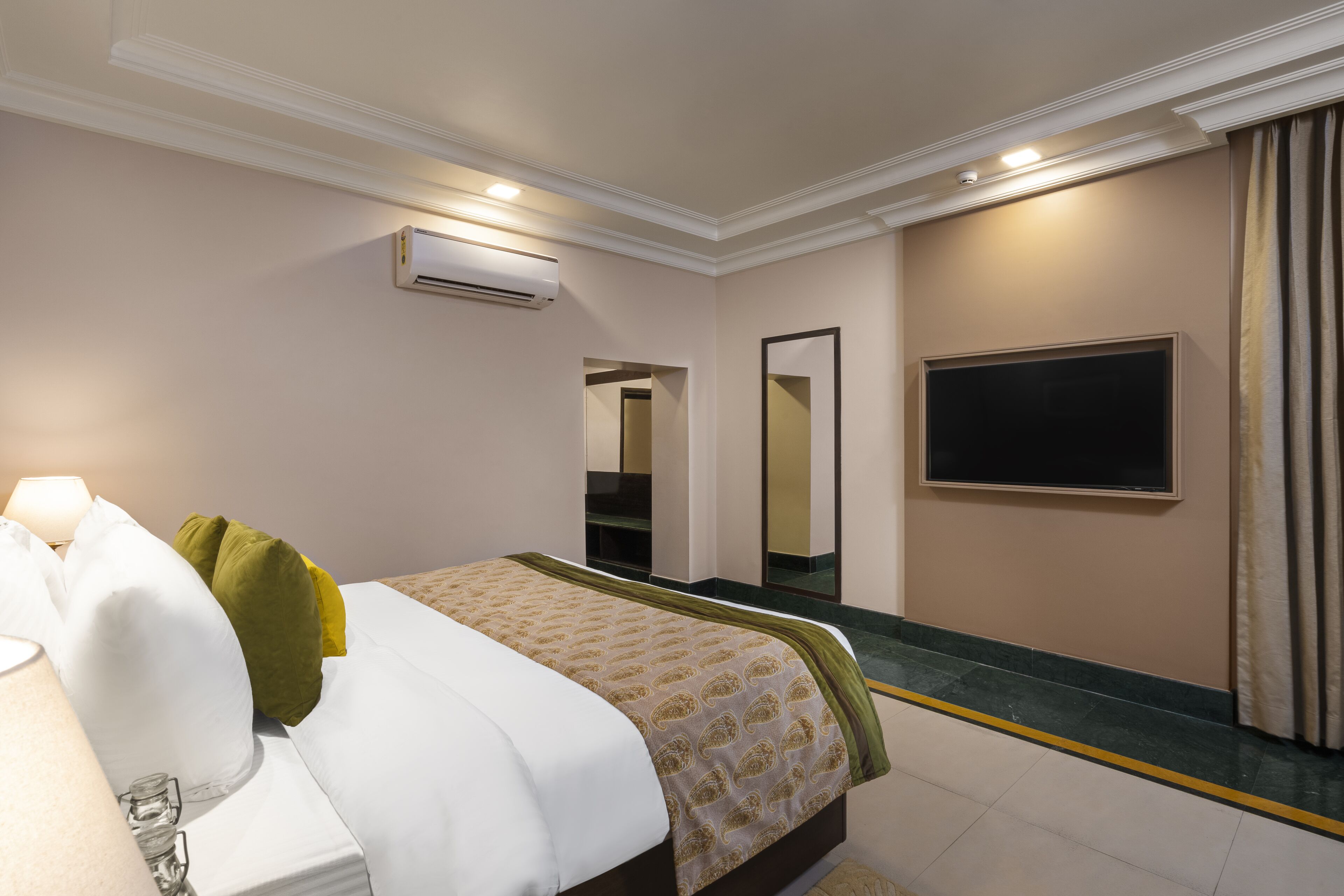 A hotel room with a bed, a bedside lamp, an air conditioning unit, a large mirror, and a television on the wall.