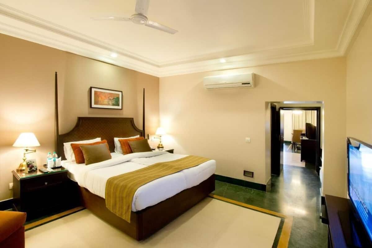 A hotel room with a bed, bedside tables, a ceiling fan, an air conditioning unit, a television, and a hallway leading to a dining area.