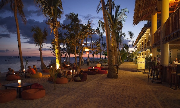 Un restaurante frente a la playa con terraza, palmeras y vistas al océano al anochecer.