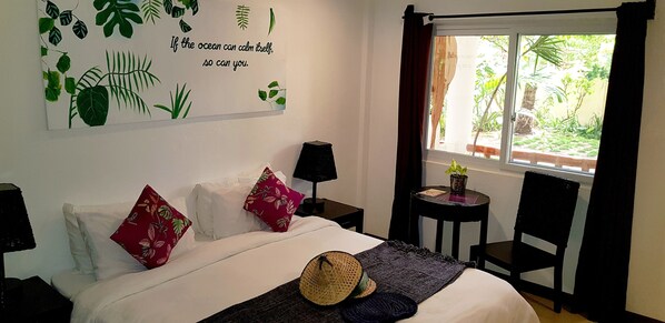A bedroom with a bed, pillows, a bedside table, a lamp, a window with curtains, and a view of greenery outside.