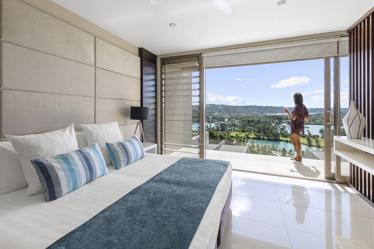 A modern hotel room with a large bed, a view of a lake and mountains, and a balcony with a person standing.