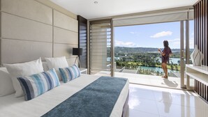 A modern hotel room with a large bed, a view of a lake and mountains, and a balcony with a person standing.