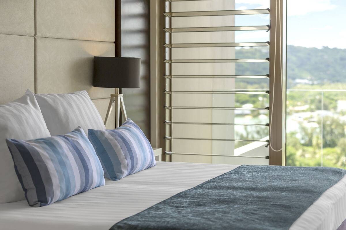 A hotel room with a bed, two pillows, a bedside lamp, and a view of greenery through a window with blinds.