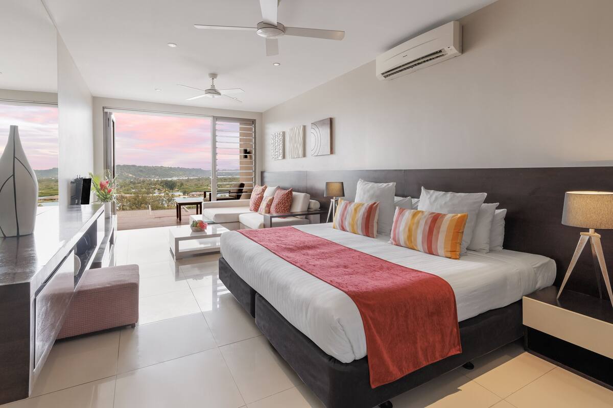 A modern hotel room with a large bed, a balcony with a city view, a ceiling fan, and an air conditioning unit.