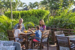 Una familia cenando al aire libre junto a una terraza de madera, rodeada de exuberante vegetación y palmeras.