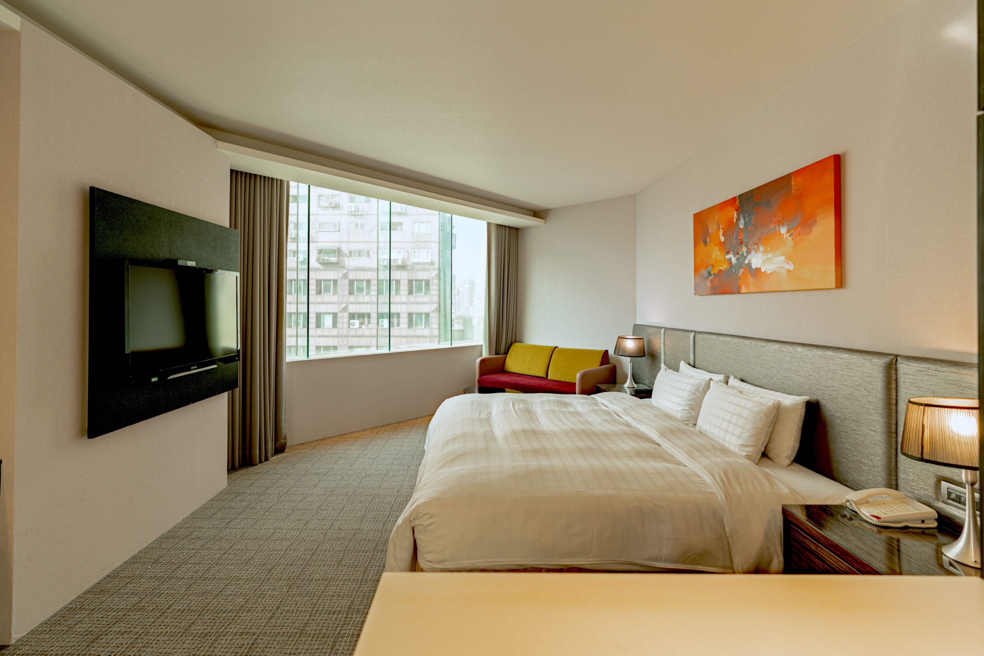 A hotel room with a bed, a TV, a sofa, a painting, and a window with a city view.