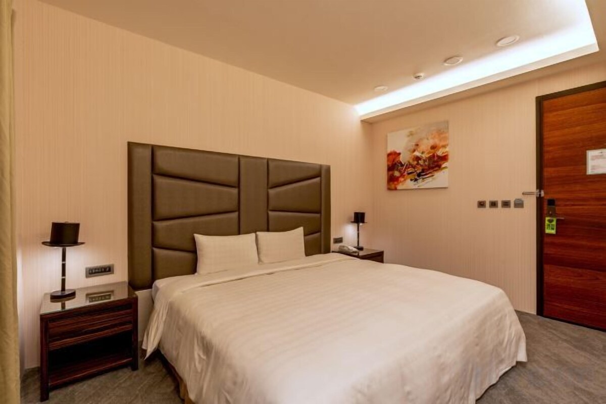 Habitación de hotel con una cama grande, mesitas de noche, una lámpara de pared y una puerta con manija.