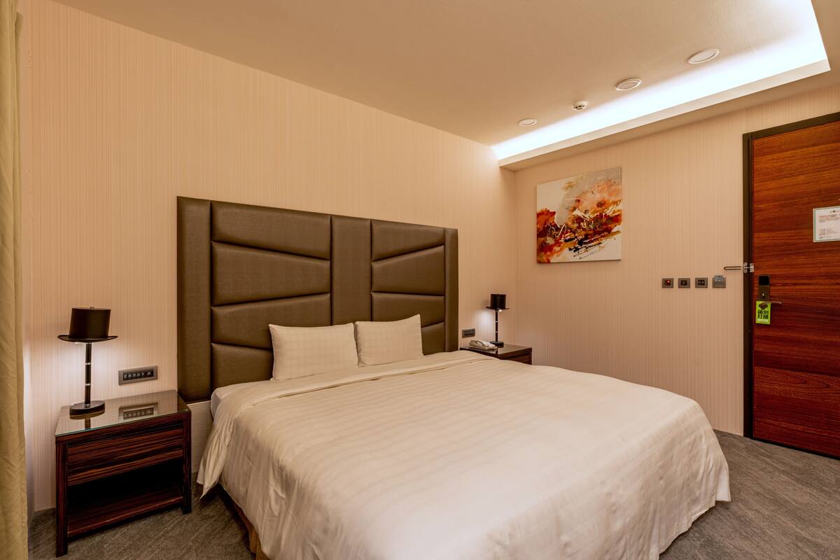 A hotel room with a large bed, a nightstand with a lamp, a wooden door, and a wall-mounted artwork.