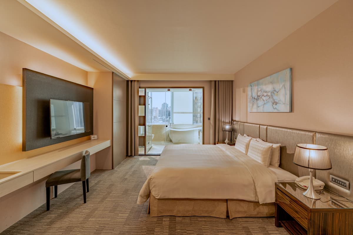 A hotel room with a bed, a desk, a chair, a TV, a bedside table with a lamp, and a painting on the wall.
