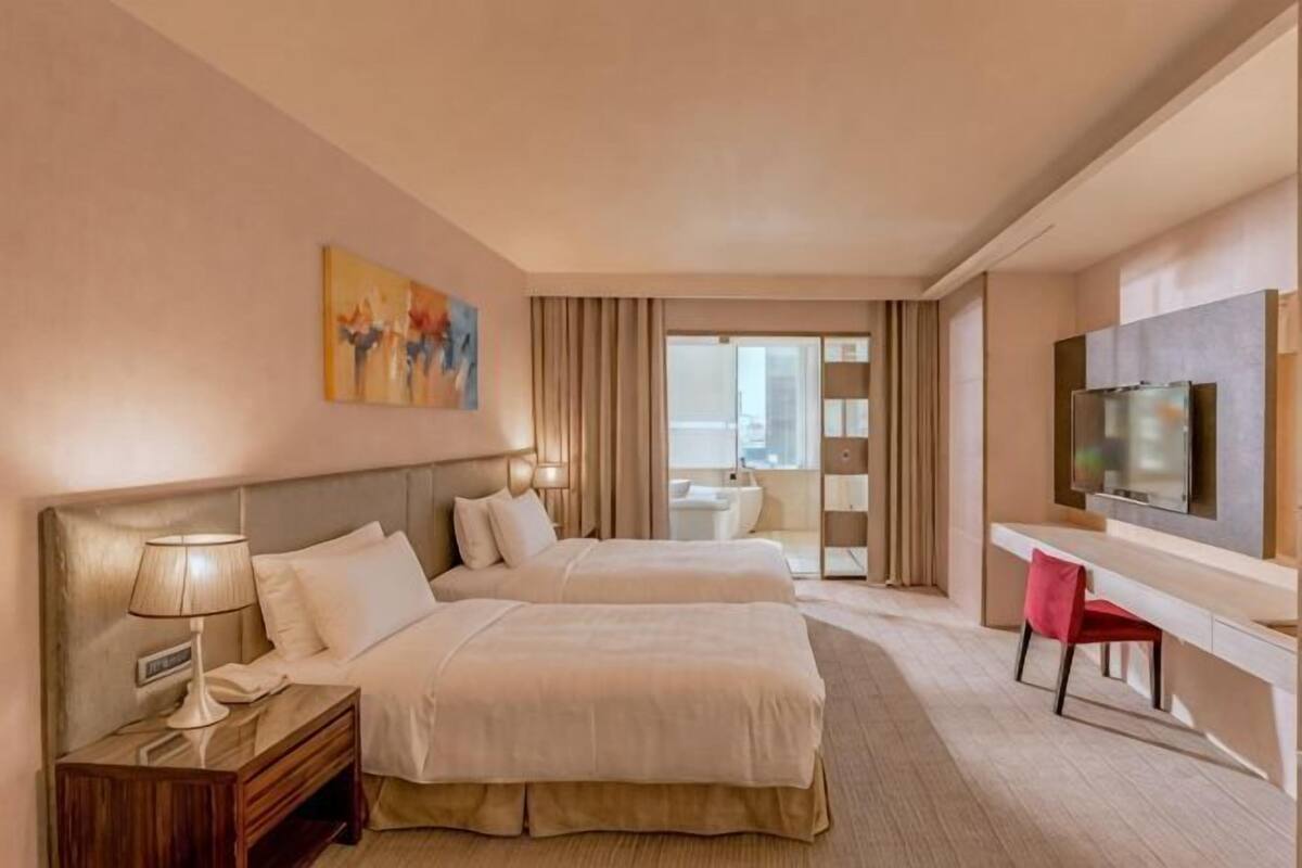 A hotel room with two beds, a desk, a TV, and a window.