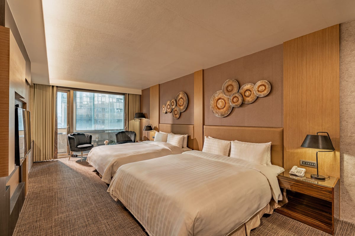 A hotel room with two beds, a desk, a chair, a lamp, and a view of the city through large windows.