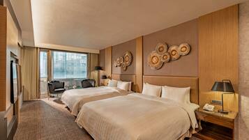 A hotel room with two beds, a desk, a chair, a lamp, and a view of the city through large windows.
