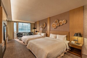 Habitación de hotel con dos camas, un escritorio, una silla, una lámpara y vistas a la ciudad a través de grandes ventanales.