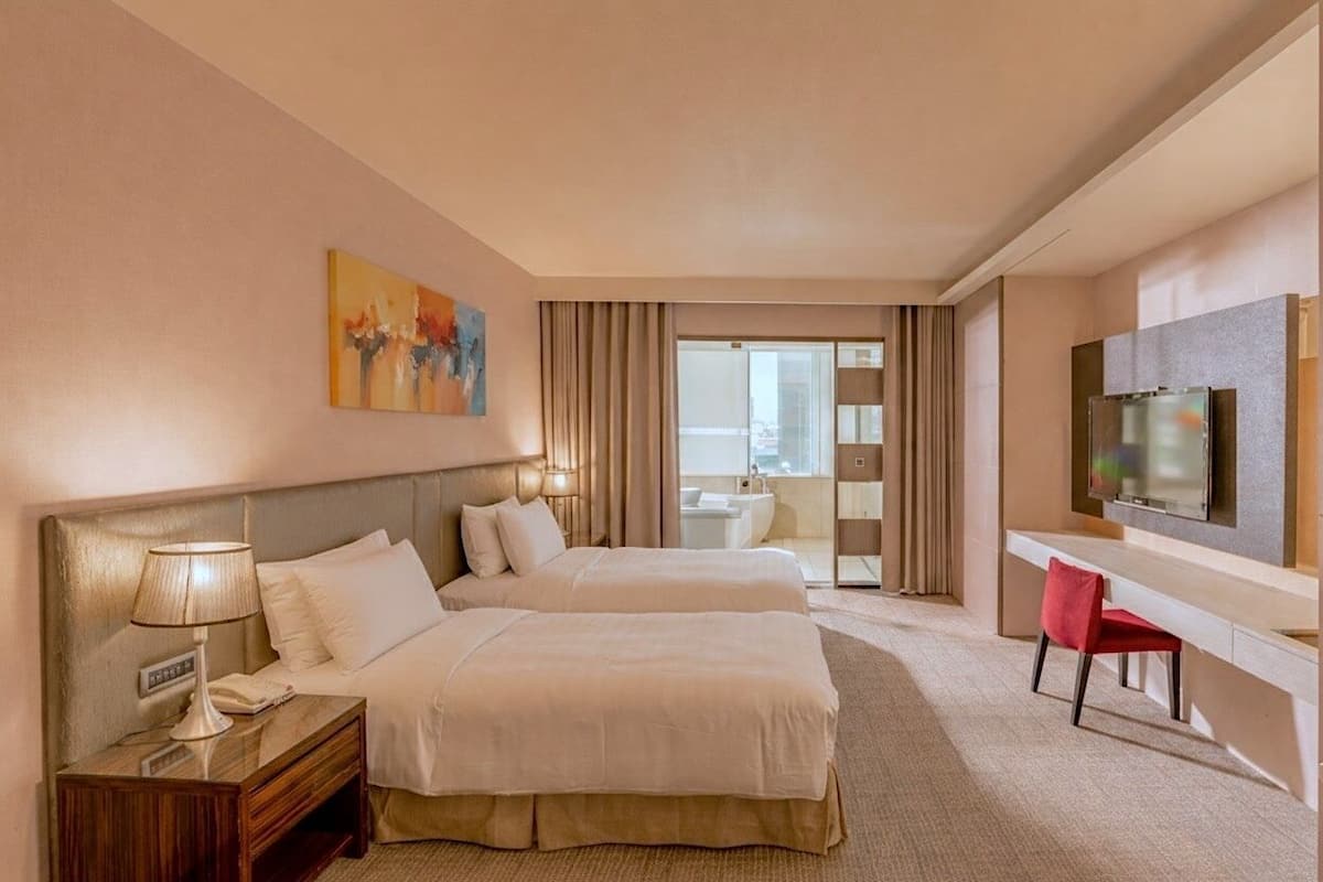 A hotel room with two beds, a desk, a TV, and a bathroom visible through a glass door.