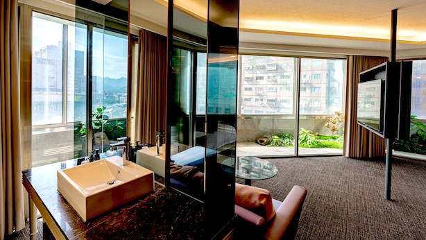 A modern hotel room with a large window, a bathroom with a sink and mirror, a flat-screen TV, and a seating area with a view of the city. - HOTEL GEIGHT TAIPEI (New Taipei City)