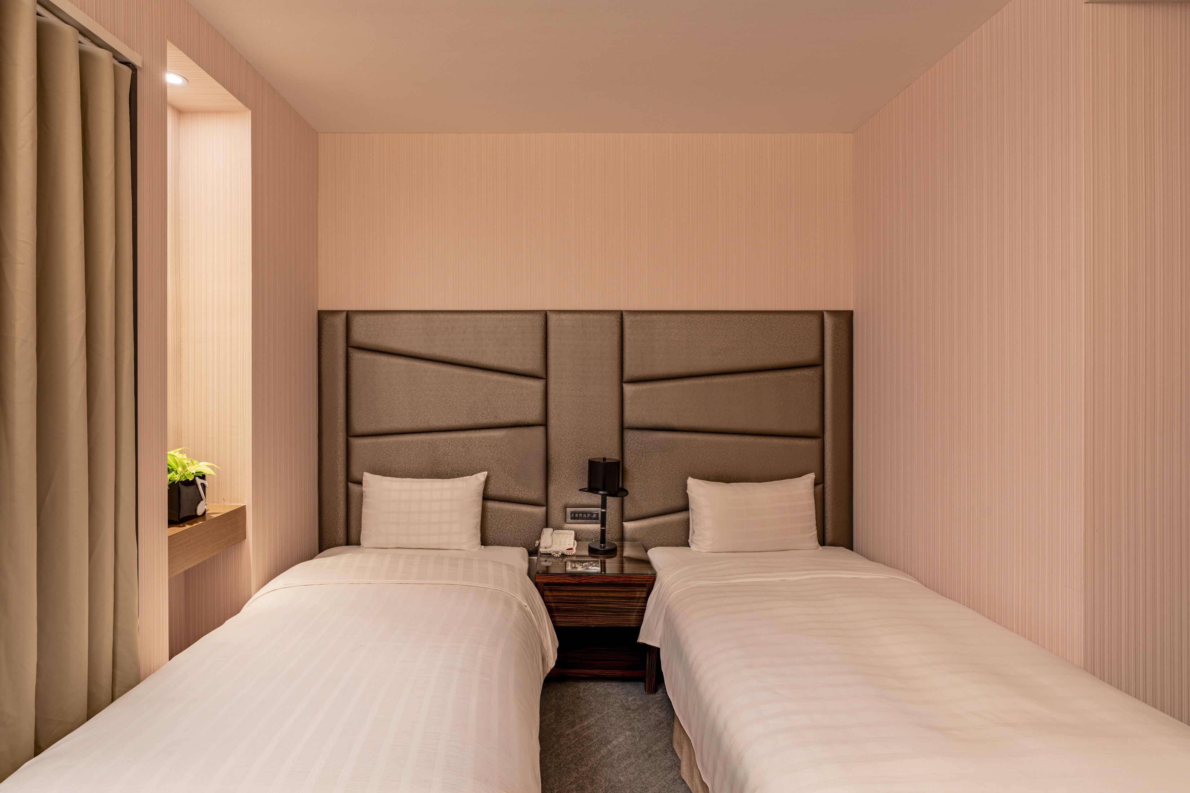 A hotel room with two beds, a bedside table with a lamp, a small plant, and a wall-mounted shelf.