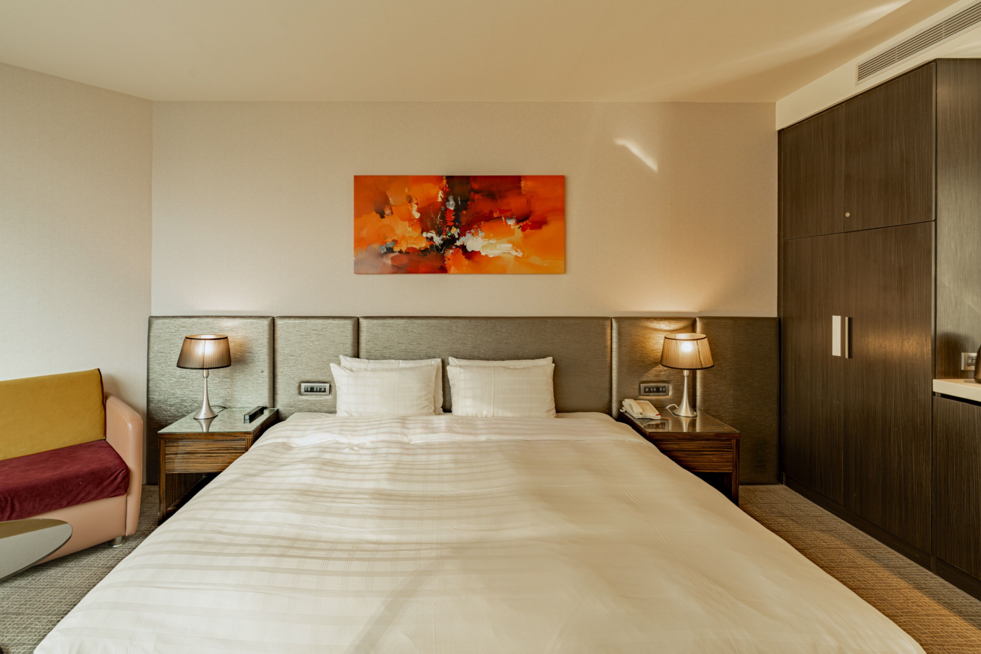 A hotel room with a large bed, two bedside tables with lamps, a wooden headboard, a red chair, and a large abstract painting on the wall.