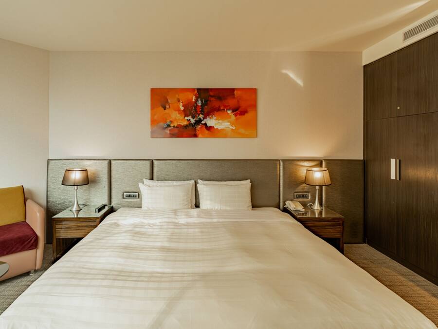 A hotel room with a large bed, two bedside tables with lamps, a wooden headboard, a red chair, and a large abstract painting on the wall.