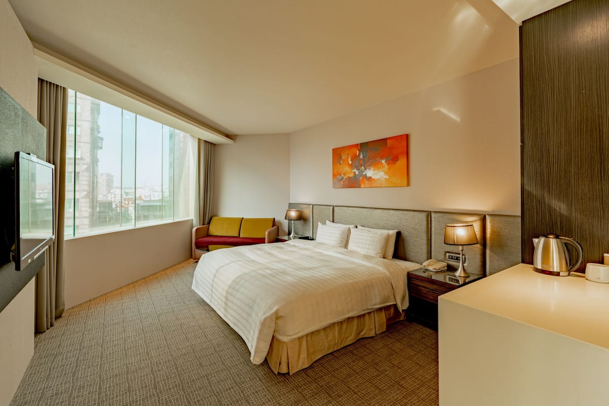 A hotel room with a large bed, a TV, a desk with a lamp, and a painting on the wall.