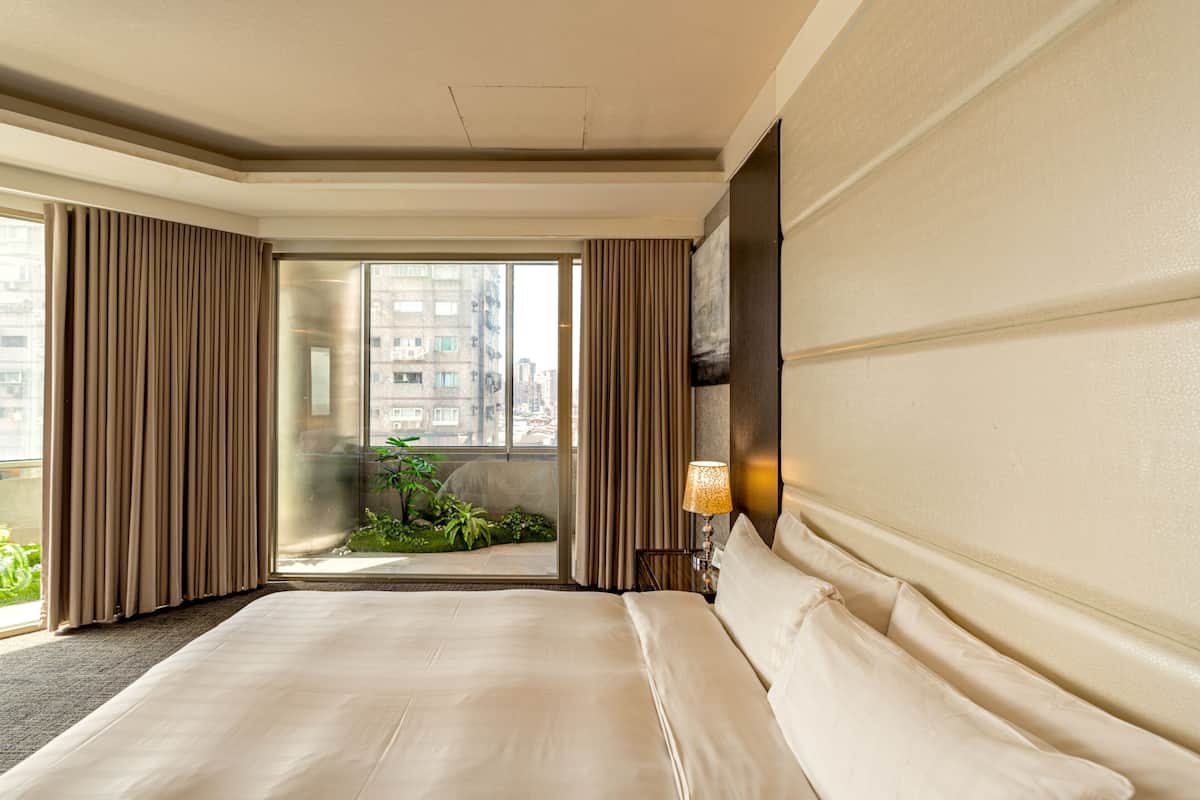 A hotel room with a large bed, a view through a glass door to an outdoor balcony, and a wall-mounted lamp.
