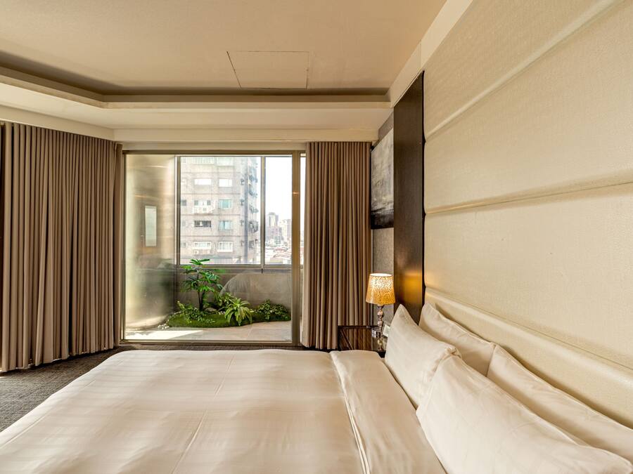 A hotel room with a large bed, a view through a glass door to an outdoor balcony, and a wall-mounted lamp.