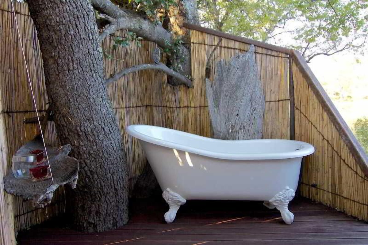 Una bañera en un entorno exterior de madera, rodeada por una cerca de bambú y un árbol.