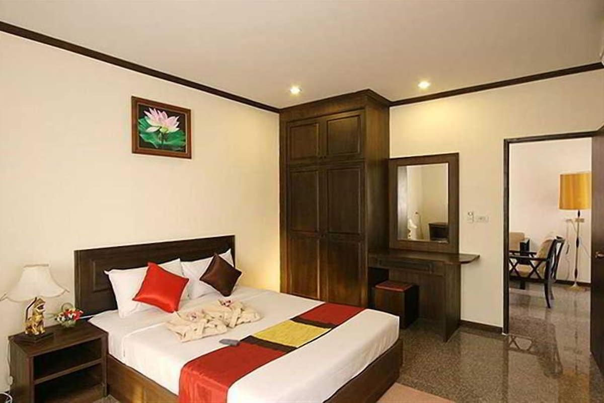 A hotel room with a large bed, a nightstand, a wooden wardrobe, a mirror, and a door leading to another room.