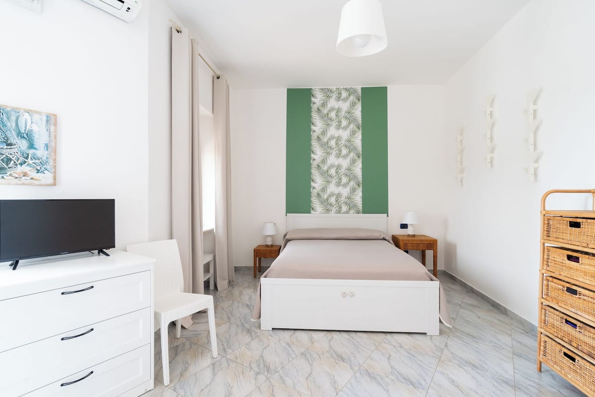 Quarto com cama, televisão em uma cômoda, cadeira, mesa de cabeceira com abajur e painel na parede com padrão de folhas.
