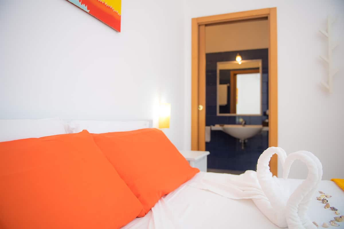Quarto de hotel com cama, almofadas laranja, toalha branca e decoração em formato de coração.