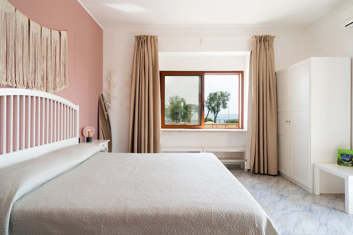 Quarto com cama, janela com cortinas, guarda-roupa e mesa de cabeceira com um livro.