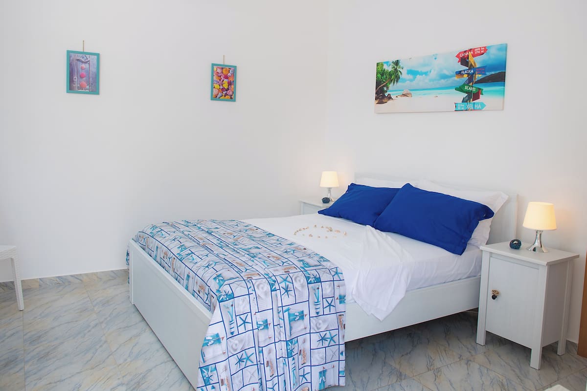 Quarto com cama, duas mesas de cabeceira, um criado-mudo, parede com três quadros e piso de mármore.