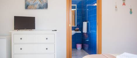 Une chambre à coucher avec un lit, une commode, un téléviseur posé sur la commode, et une salle de bain visible à travers une porte ouverte.