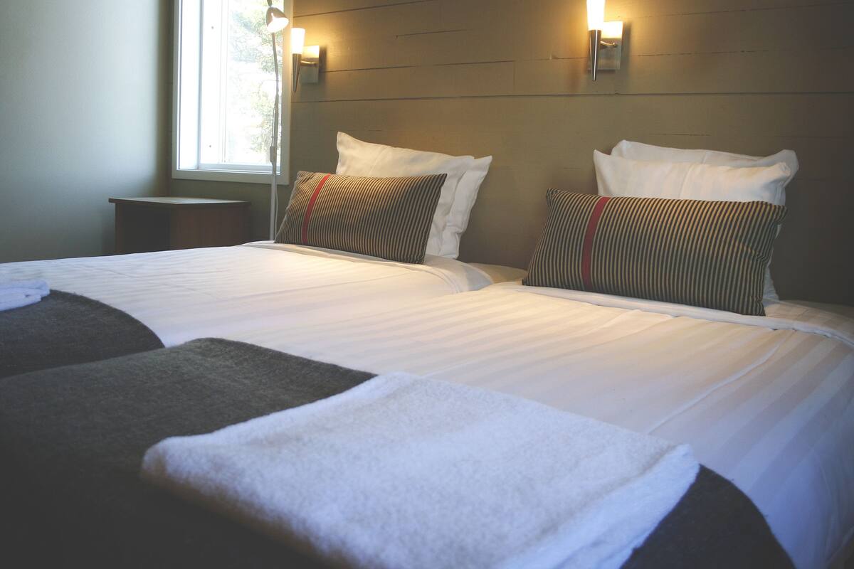A hotel room with two beds, each with white linens and striped pillows. There is a small bedside table and a window with a view of trees.