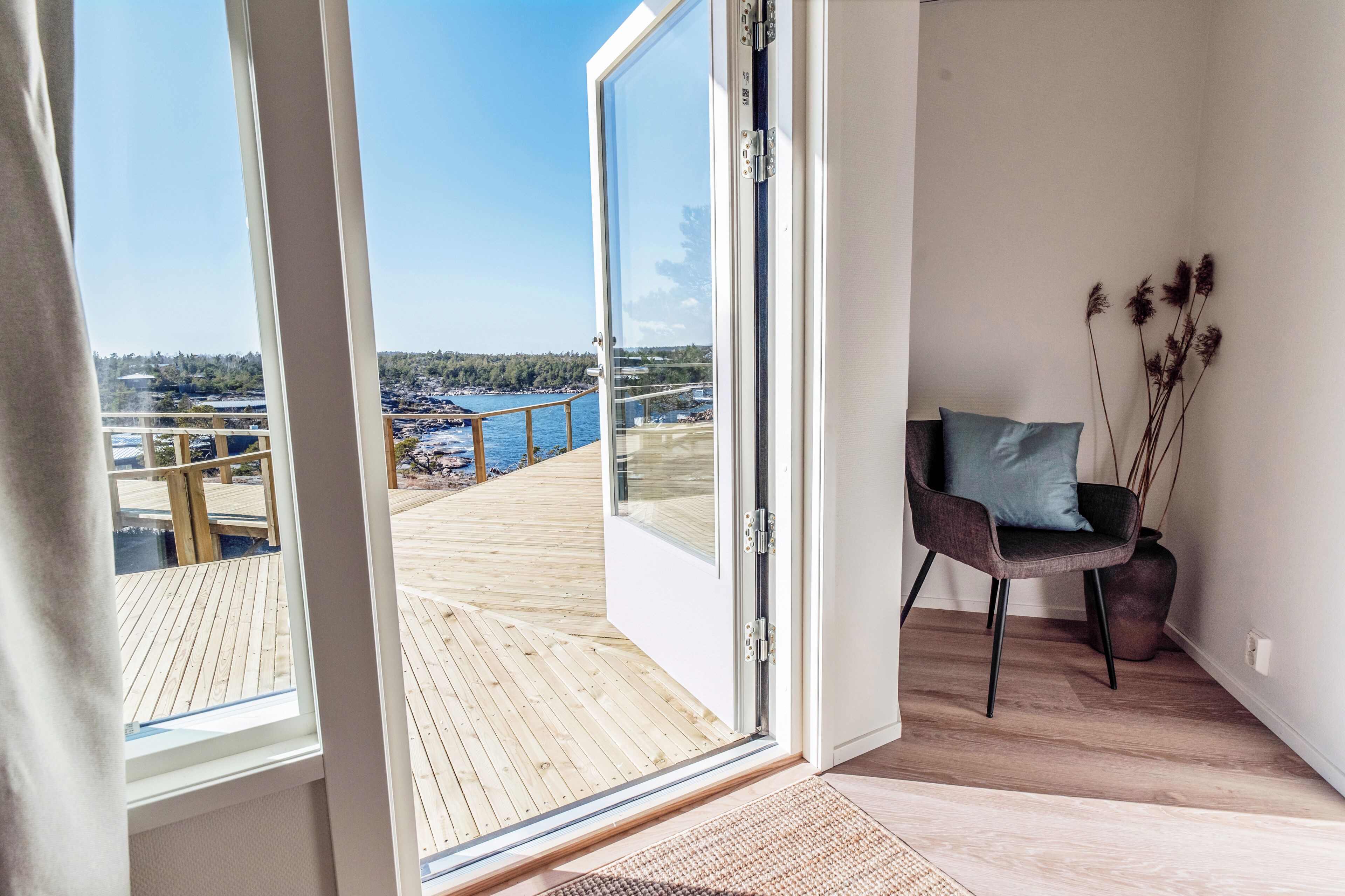 A room with a view of a wooden deck and water, a chair with a cushion, and a potted plant.