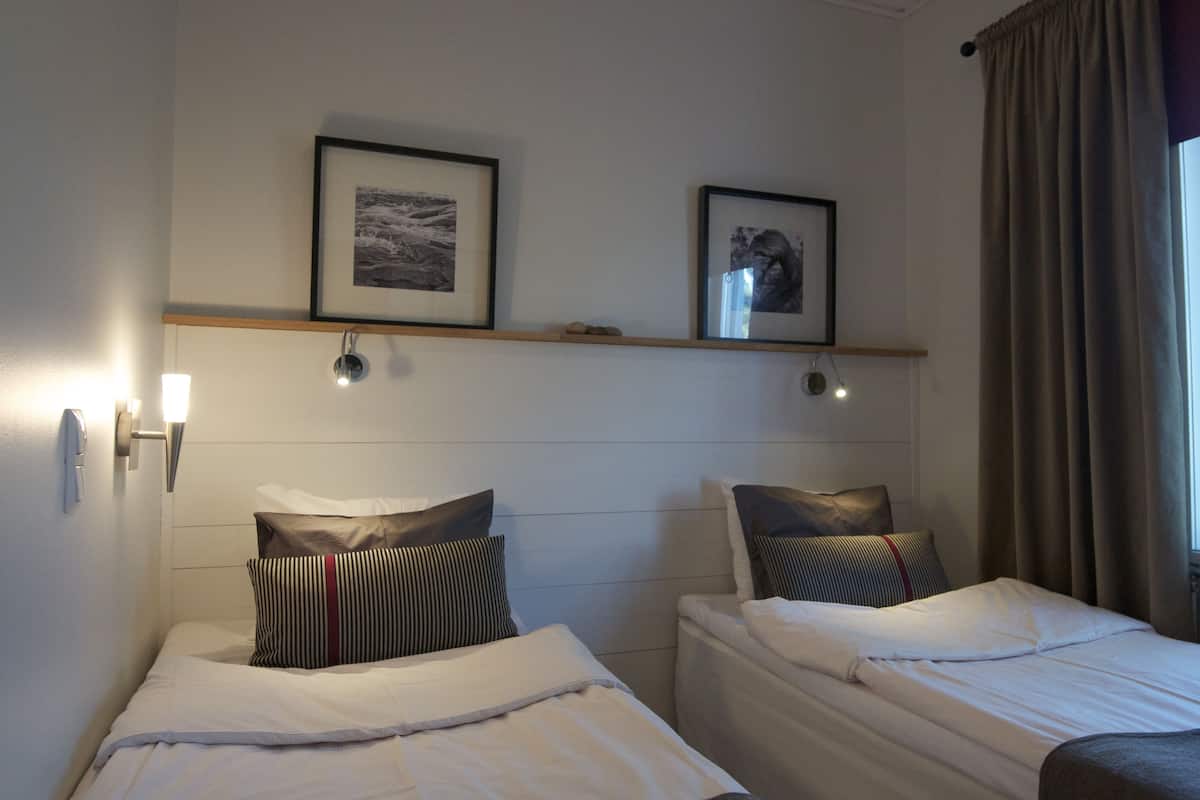 A hotel room with two single beds, a wooden headboard, wall-mounted light fixtures, and framed artwork above the beds.