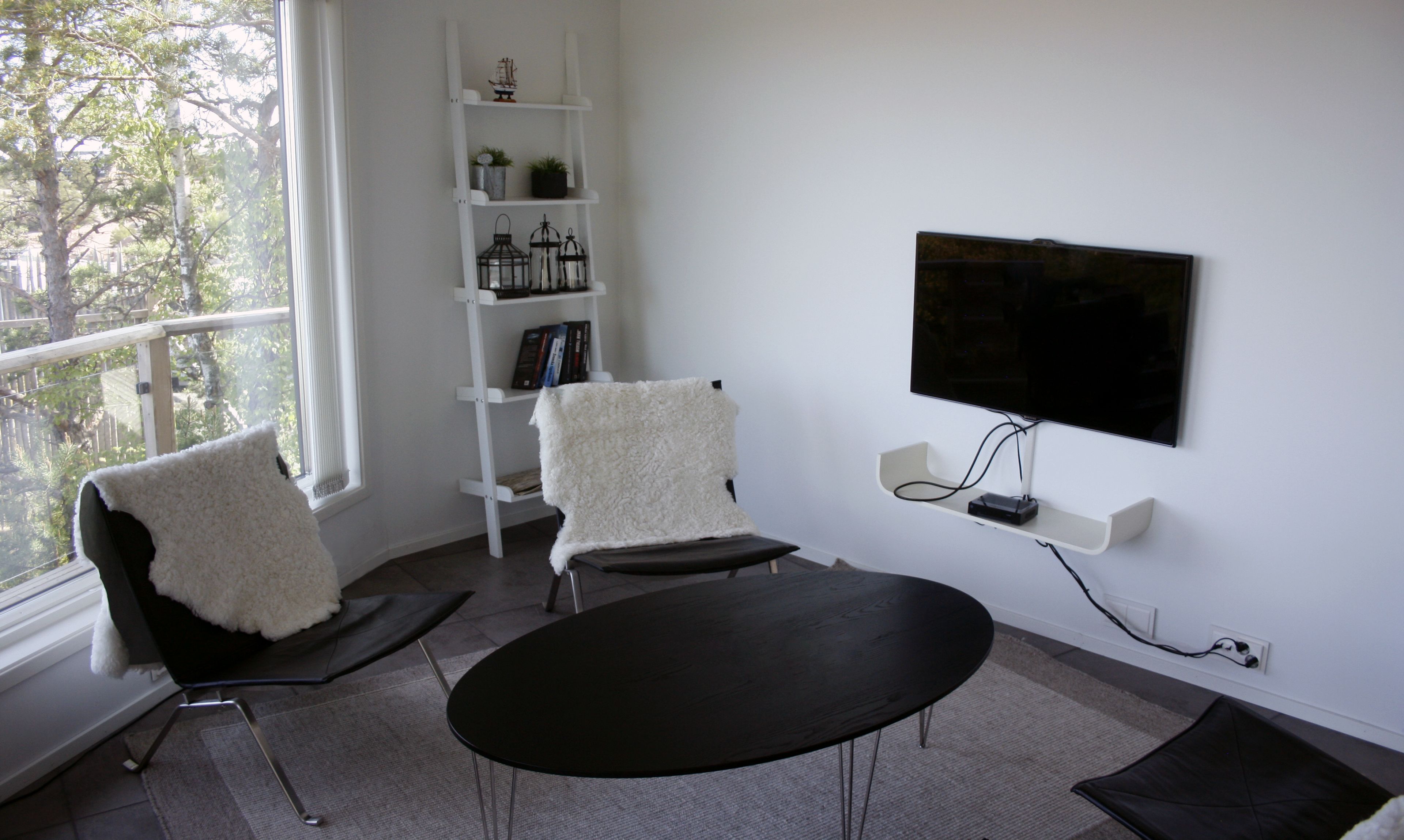 A modern living room with a black round table, two black chairs, a white bookshelf, and a flat-screen TV mounted on the wall.