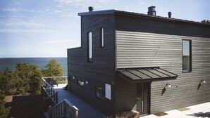 A modern two-story building with a deck overlooking the sea, featuring a minimalist design and large windows.