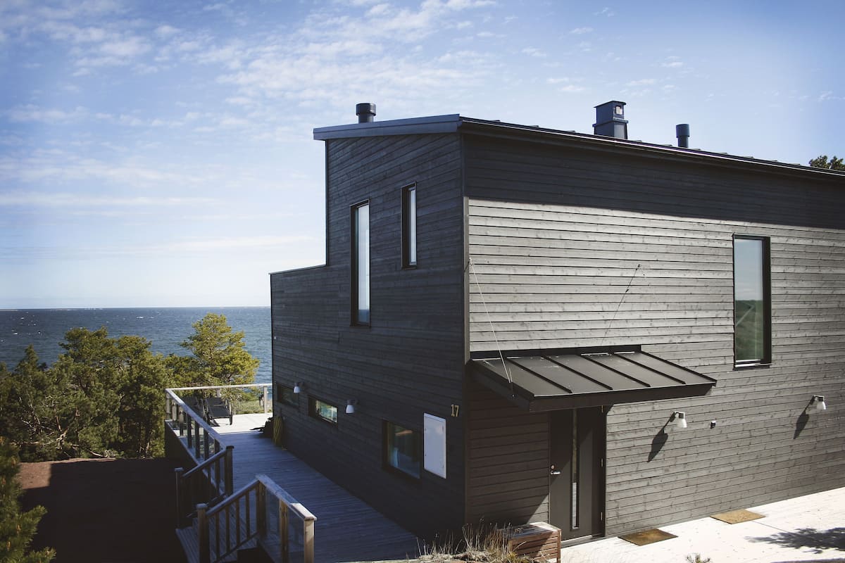 A modern two-story building with a deck overlooking the sea, featuring a minimalist design and large windows.