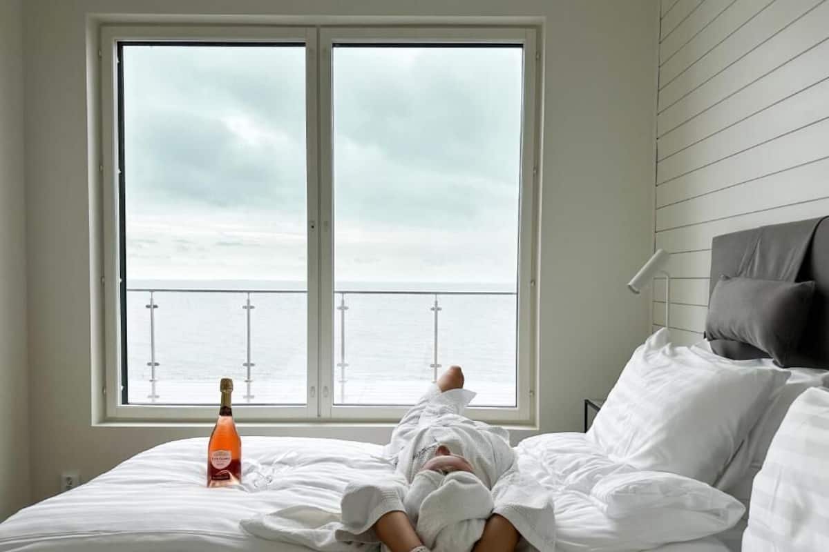 A bed with a bottle of champagne, a view of the sea through a window, and a grey pillow.