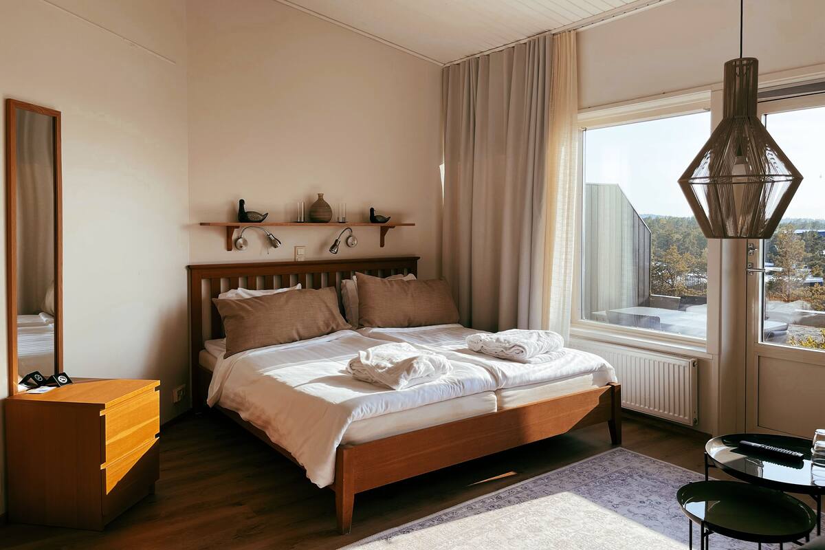 A bedroom with a wooden bed, a wooden dresser, a bedside table, a hanging lamp, a window with curtains, and a view of the outside.