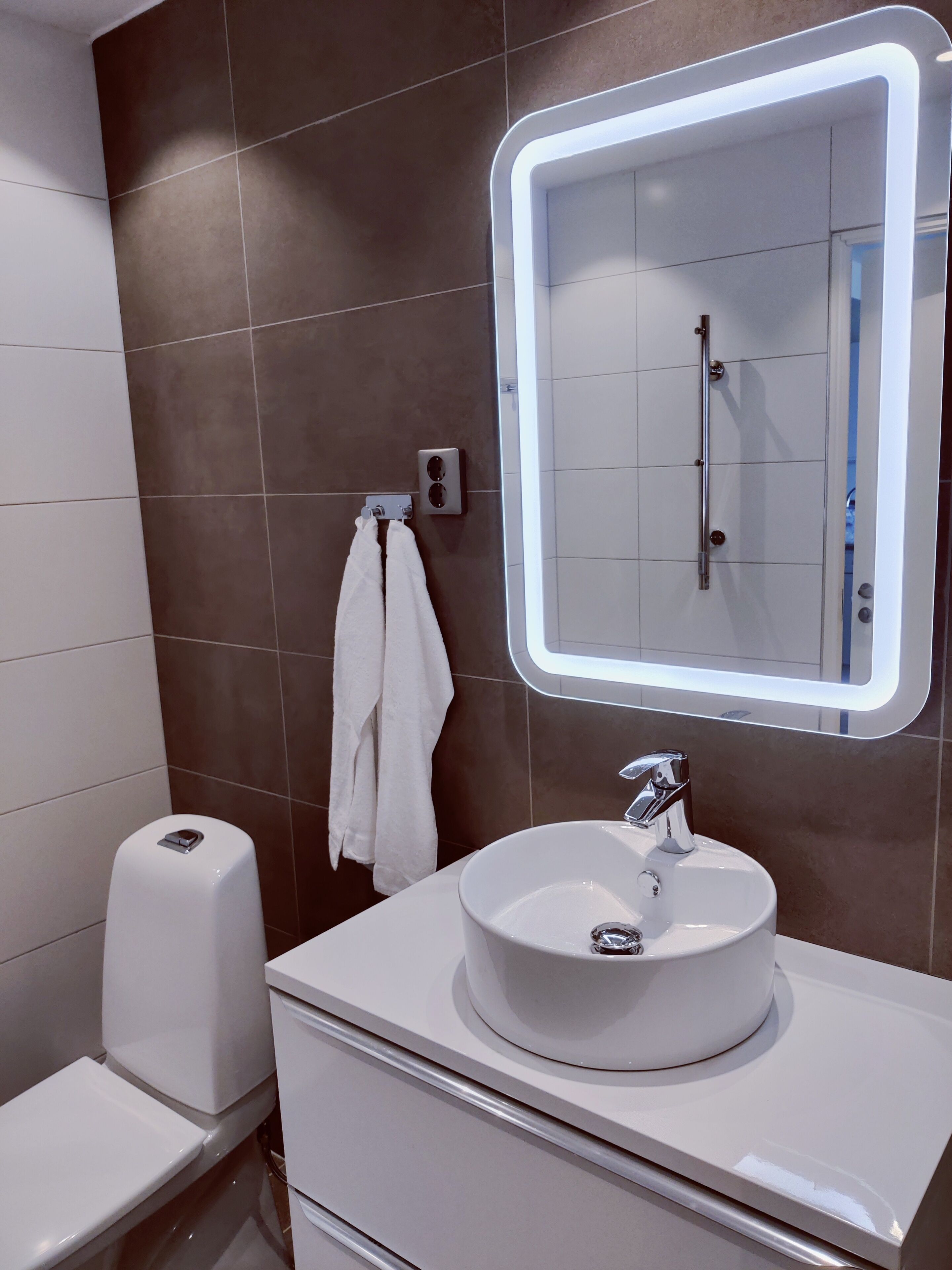 A modern bathroom with a white sink, a mirror with a light, a towel hanging on the wall, and a toilet.