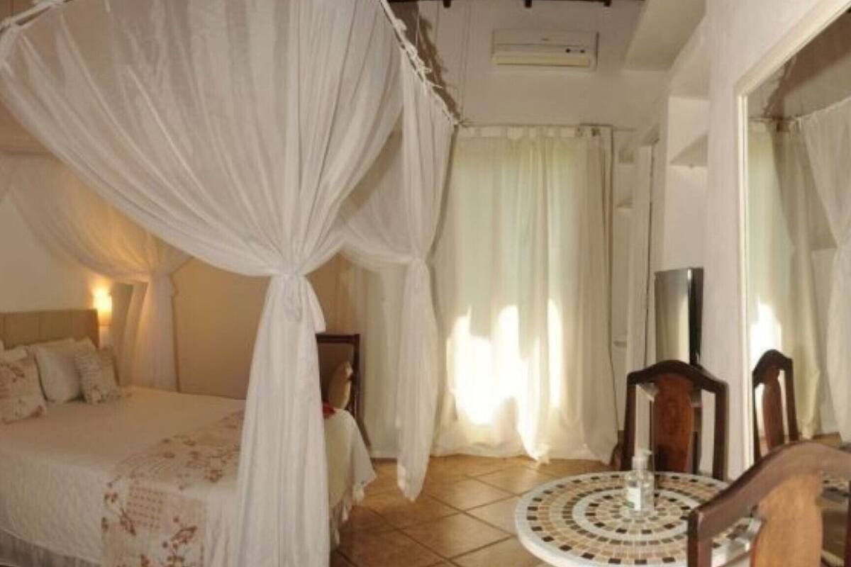 Un dormitorio con una cama con dosel, una mesita de noche, una mesa redonda con sillas y una ventana con cortinas.