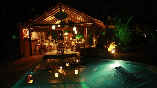 A nighttime view of a poolside area with a thatched-roof structure, lounge chairs, and a pool reflecting lights. - Pousada Magia Verde (Paraty)