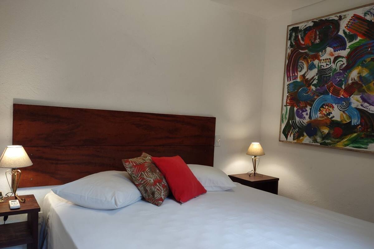 Habitación de hotel con cabecera de madera, cama con ropa blanca, almohadas rojas y estampadas, dos mesitas de noche con lámparas y un cuadro abstracto colorido en la pared.